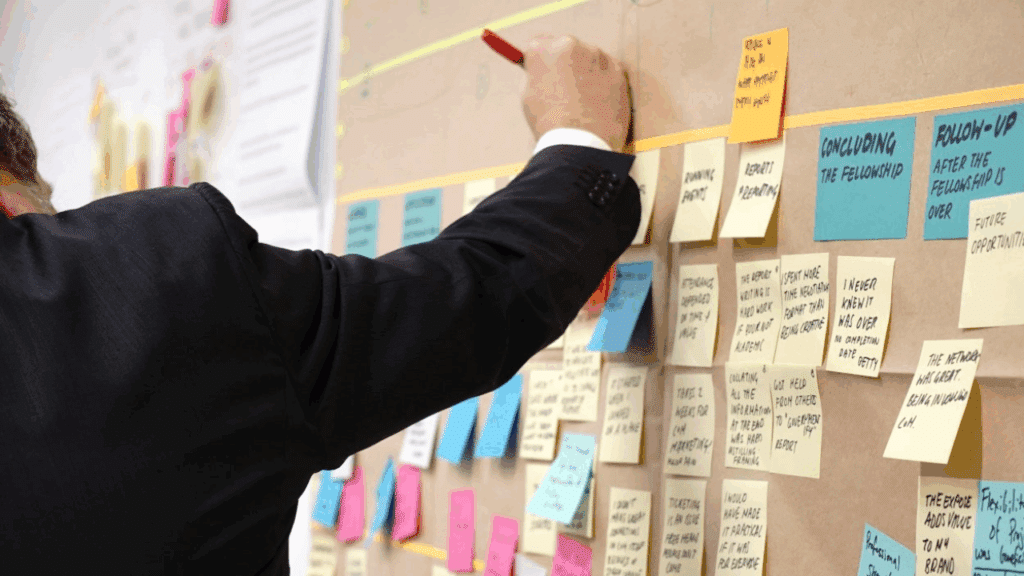 um homem utiliza um organizador de tarefas físico em sua parede com diversos post it colados.